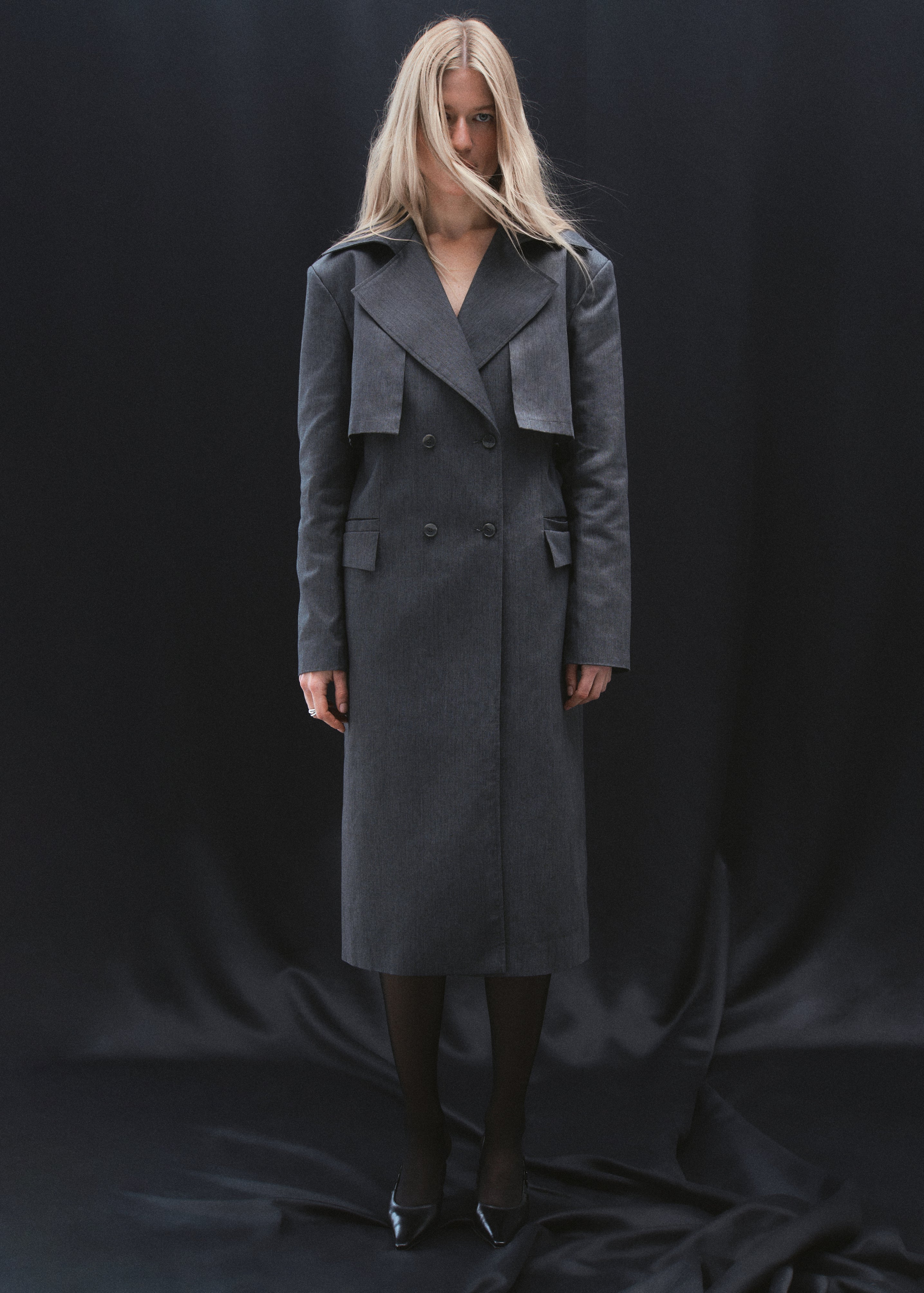 A model standing in a dark grey trench coat with four front buttons, created from recycled polyester and organic cotton blend