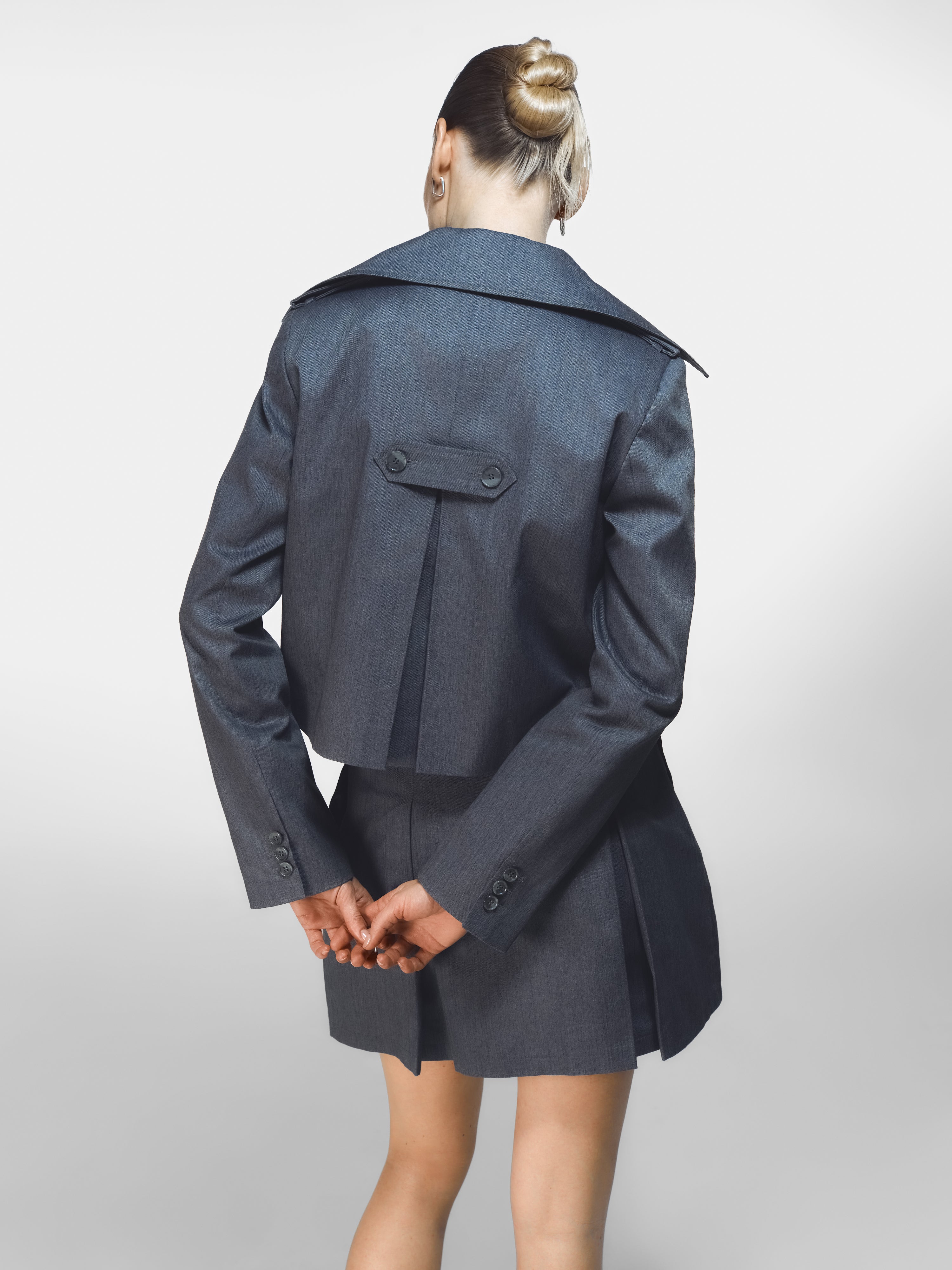 A model posing showing the back details of the SeaSycle jacket and mini skirt by HER CIPHER in color Graffite