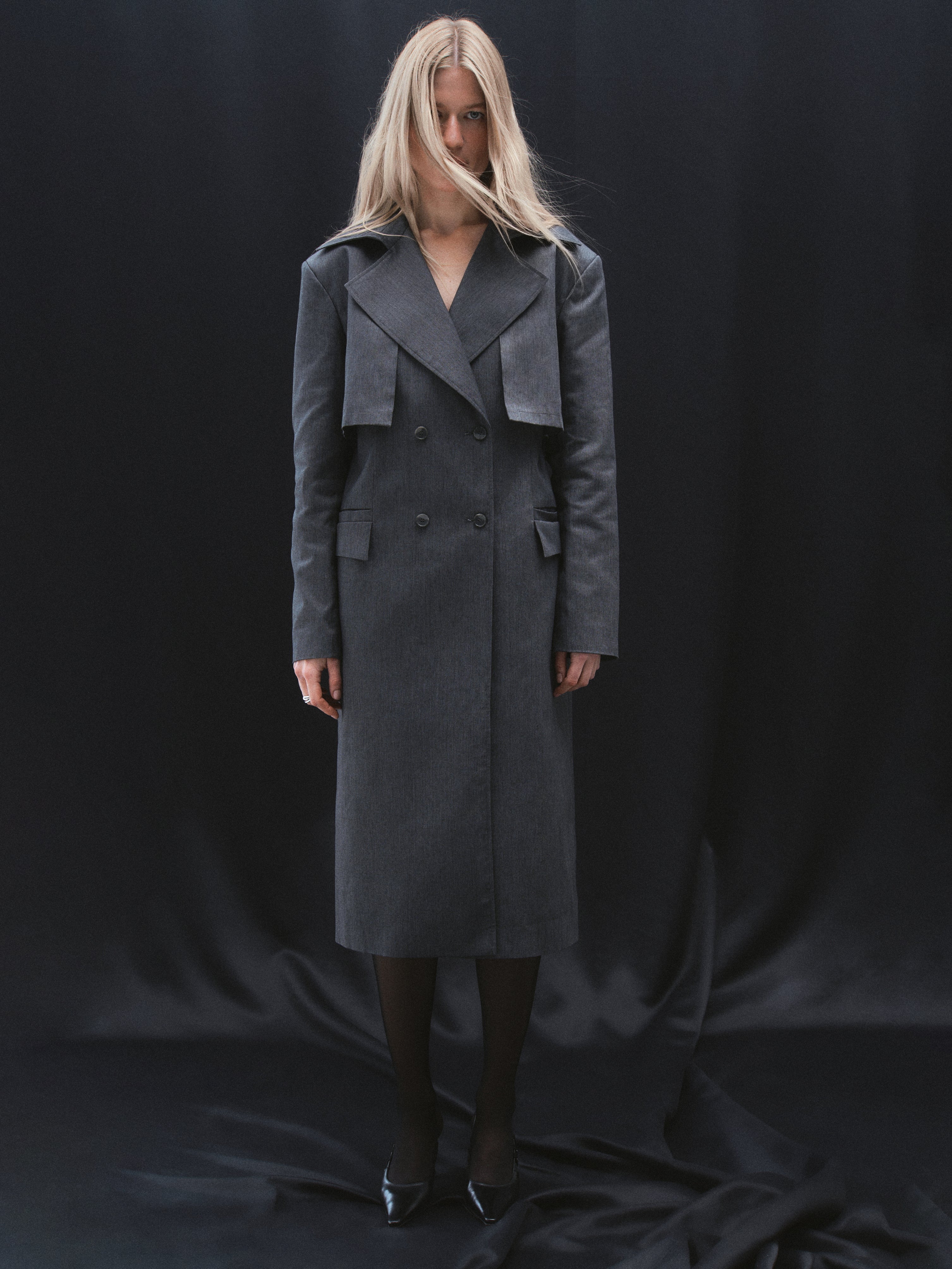 A model standing in a dark grey trench coat with four front buttons, created from recycled polyester and organic cotton blend