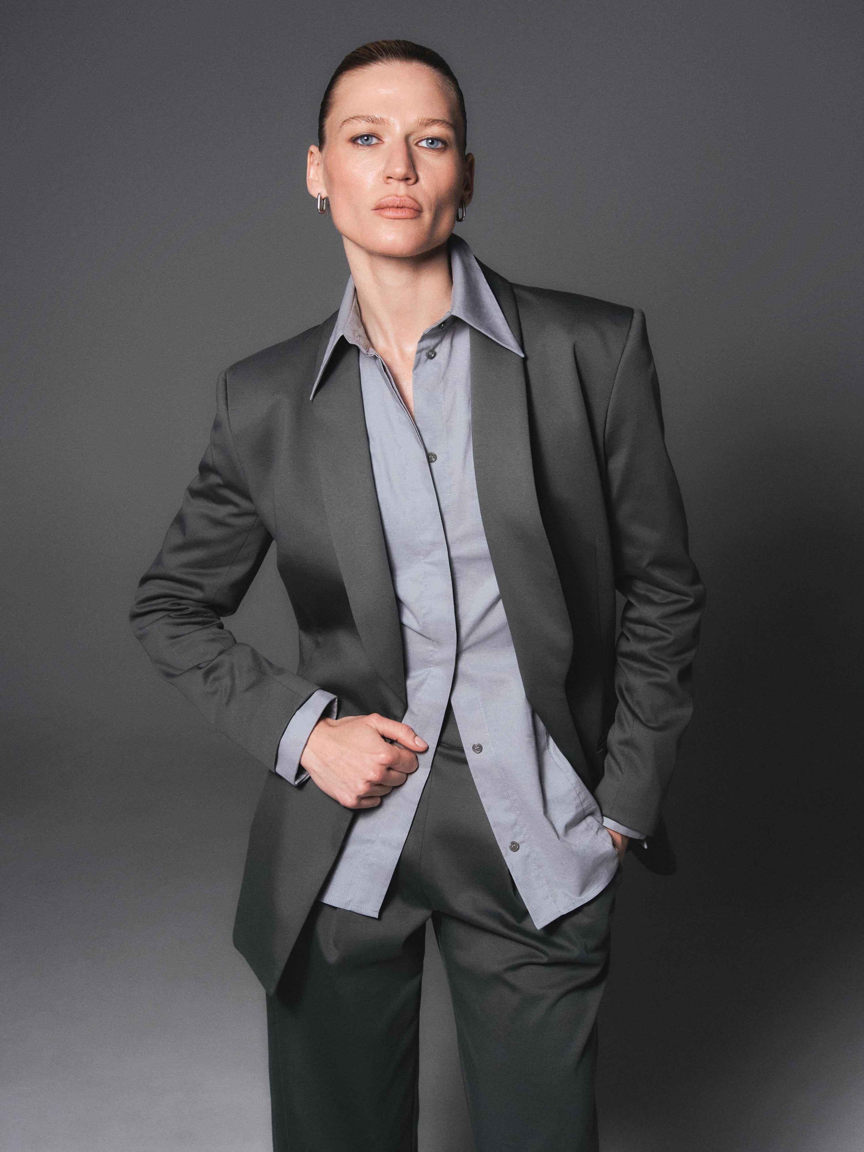 A model posing wearing an organic cotton outfit, that consists of an organic interlock blazer and pants and organic cotton shirt in grey color